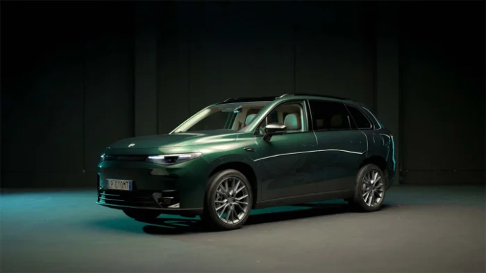 Ein dunkelgrüner SUV steht in einem Raum mit dunklem Hintergrund. Das Fahrzeug ist von der Seite zu sehen, mit der Front leicht zum Betrachter gedreht. Die Scheinwerfer sind eingeschaltet und reflektieren Licht. Das Auto hat ein modernes, elegantes Design und steht auf silbernen Felgen.