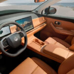 Innenraum eines Autos mit orangefarbenen Ledersitzen und -verkleidung. Ein modernes Lenkrad mit digitalen Anzeigen und ein großer Bildschirm in der Mitte des Armaturenbretts sind deutlich sichtbar. Das Auto steht am Strand, mit Blick auf das Meer und eine Küstenlandschaft im Hintergrund.