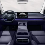 Innenraum eines Autos mit modernem Design. Der Fokus liegt auf dem Armaturenbrett mit einem großen Display auf der rechten Seite des Lenkrads. Die Sitze und Innenverkleidungen sind in einem dunklen lila Farbton gehalten. Eine blaue LED-Leiste verläuft entlang des Armaturenbretts. Im Hintergrund ist der