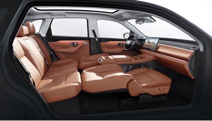 Innenraum eines Autos mit braunen Ledersitzen. Die Rücksitze sind durchgehend und bieten Platz für drei Personen. Vordersitze sind stark zurückgelehnt, um zusätzlichen Komfort zu zeigen. Die Mittelkonsole hat einen Bildschirm und verschiedene Bedienelemente. Große Fenster ermöglichen viel natürliches Licht.