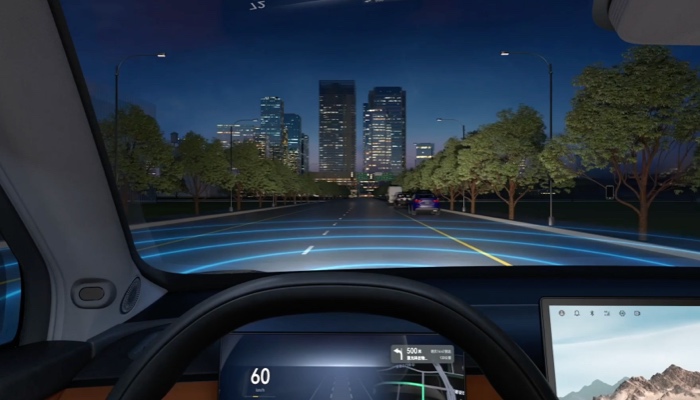 Das Bild zeigt das Innere eines Fahrzeugs bei Nacht. Aus der Perspektive des Fahrers ist eine beleuchtete Straße mit mehreren Fahrspuren zu sehen, die in Richtung einer Stadt mit beleuchteten Hochhäusern führt. Auf dem Armaturenbrett des Fahrzeugs sind digitale Anzeigen zu erkennen. Bäume säumen die Straße